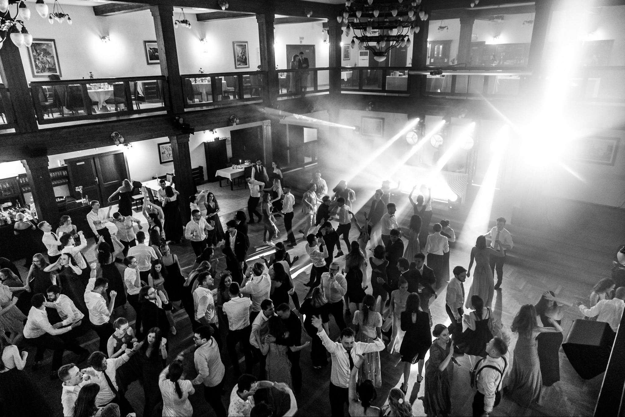 Czarno-białe zdjęcie z góry przedstawiające bawiących się ludzi na parkiecie podczas wydarzenia, oświetlonych reflektorami i z widocznym dymem, z balkonem i stołami w tle.