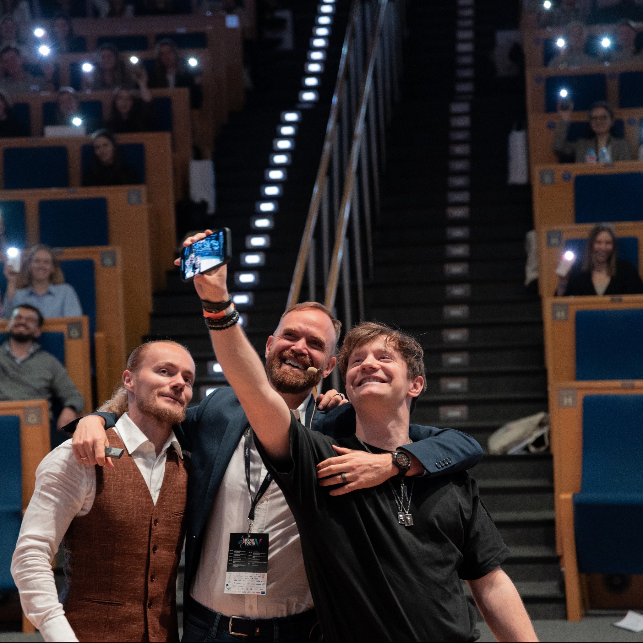 Trzech uśmiechniętych mężczyzn robi sobie selfie na tle widowni z podniesionymi telefonami.