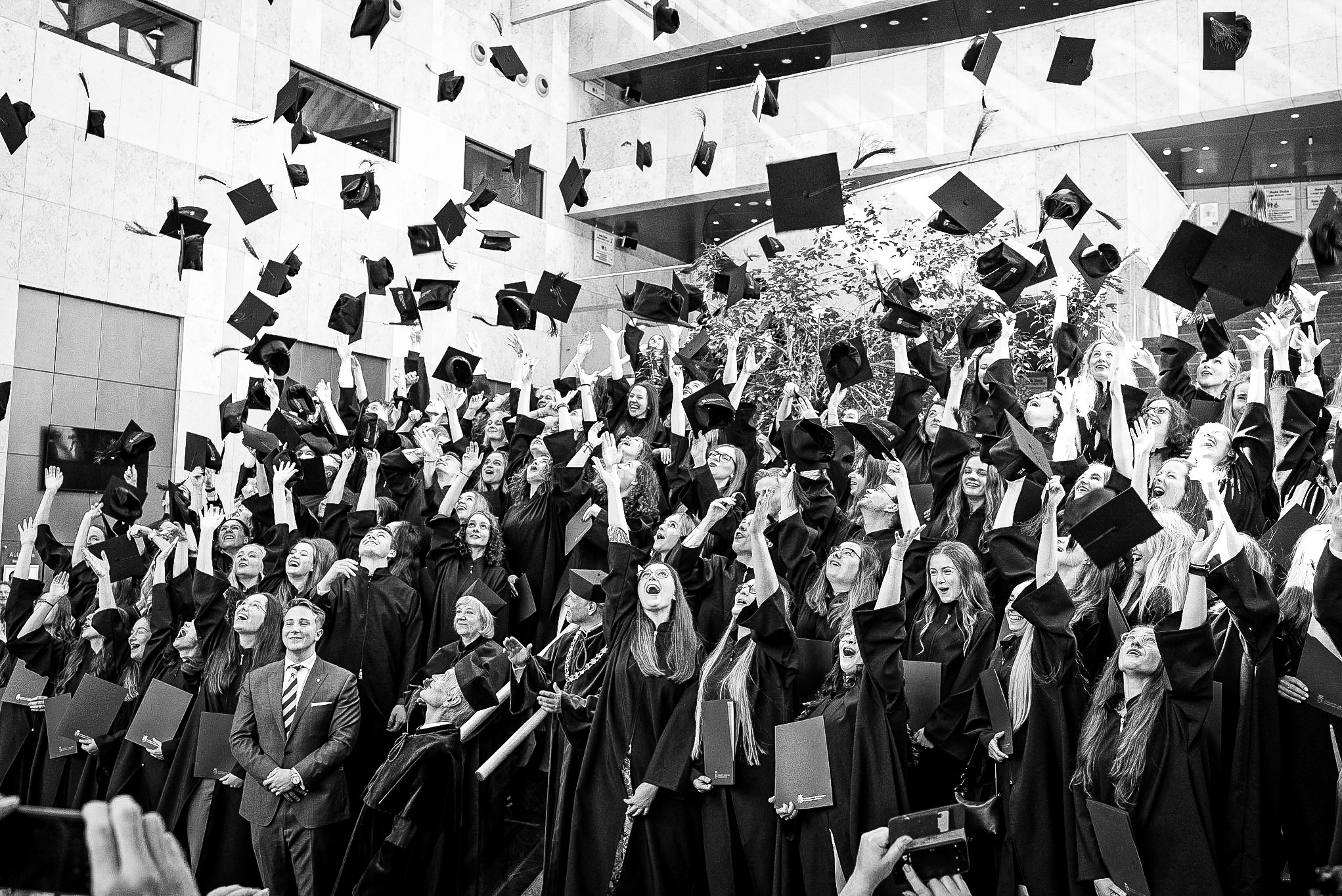 Czarno-białe zdjęcie grupy absolwentów w togach wyrzucających w górę birety, z uśmiechniętym mężczyzną w garniturze na pierwszym planie, w tle budynek uczelni z drzewami.