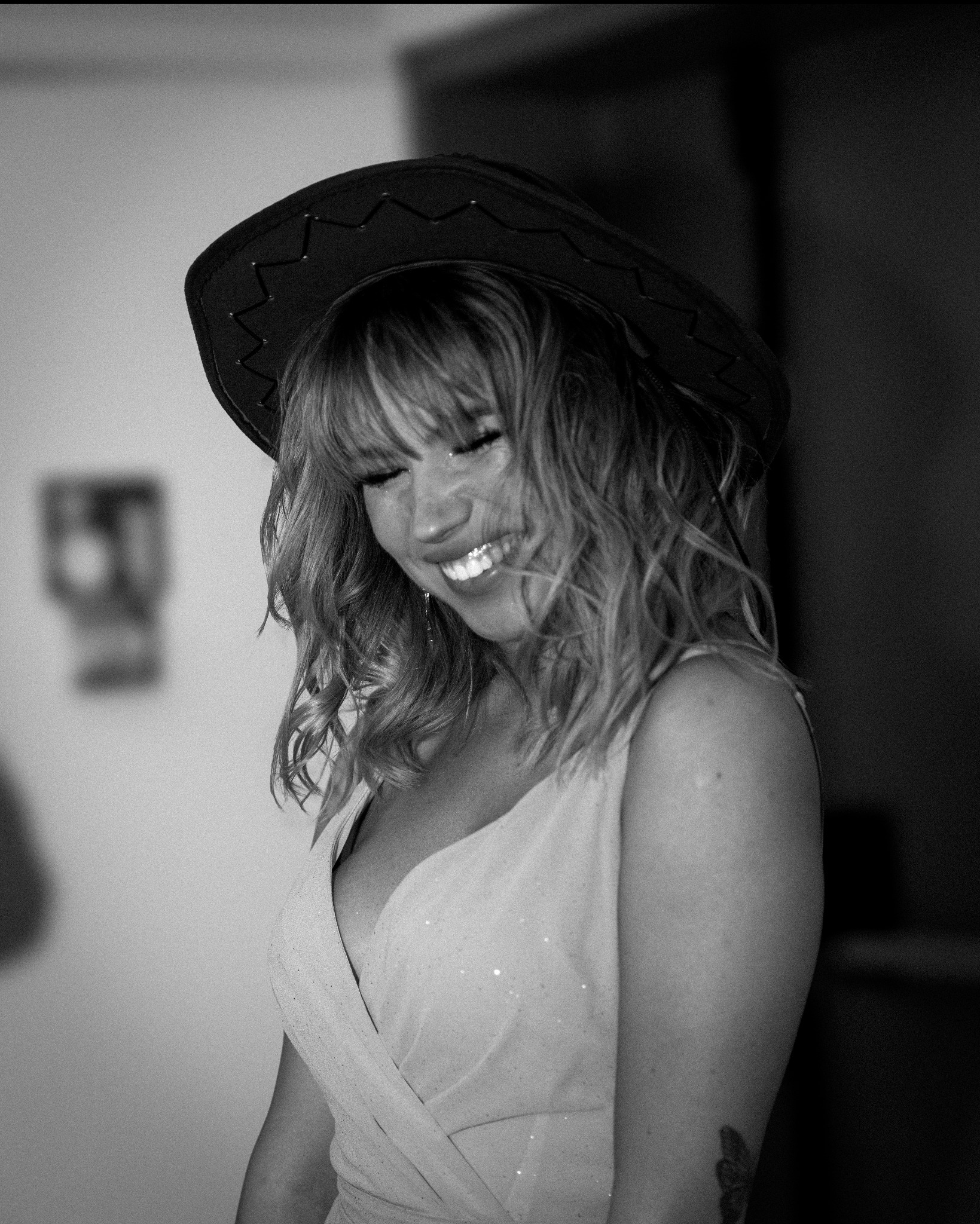 Portret uśmiechniętej kobiety w kowbojskim kapeluszu i sukience, czarno-białe zdjęcie z widocznym tatuażem na ramieniu.