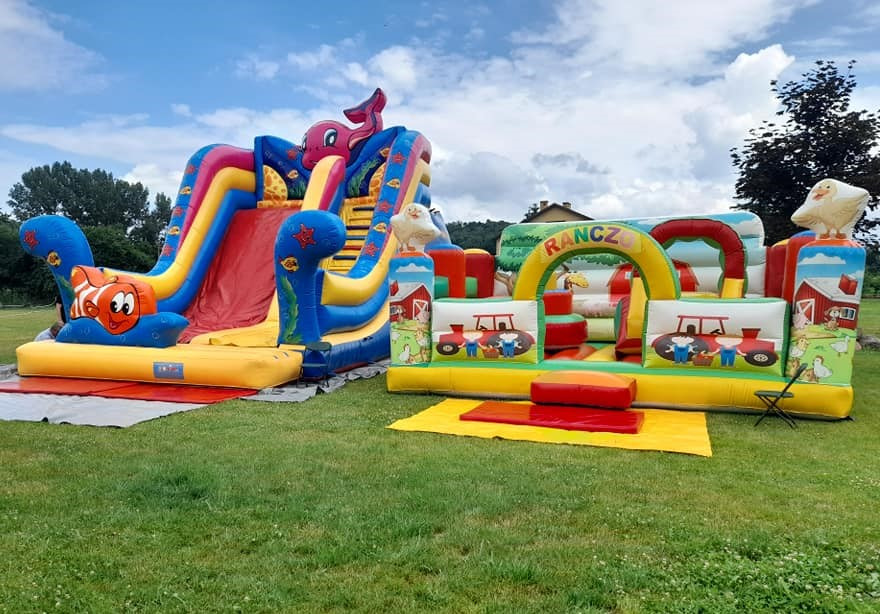Dwa kolorowe dmuchane zamki na trawie: jeden ze zjeżdżalnią z motywem morskim (ryba błazenek i ośmiornica), drugi z napisem 'RANCZ' i motywami wiejskimi (traktory, kury, stodoła).