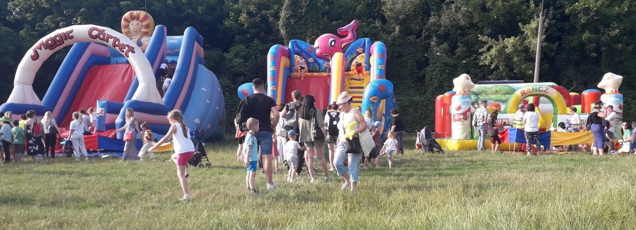 Rozległy teren zielony z kilkoma kolorowymi dmuchanymi zamkami i zjeżdżalniami, wokół których zgromadzeni są ludzie, w tym dzieci, w słoneczny dzień.