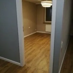 Puste pomieszczenie po remoncie: świeżo ułożona podłoga z paneli w odcieniach drewna, szare ściany, widoczny fragment okna z grzejnikiem i nowoczesna lampa sufitowa. Widok z perspektywy wejścia...