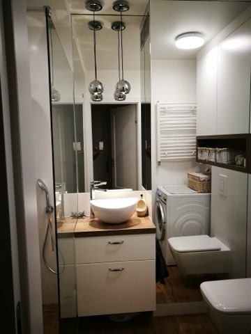 Nowoczesna, jasna łazienka z umywalką nablatową w kształcie misy, drewnianym blatem, białą szafką, pralką i wiszącą toaletą, oświetlona trzema chromowanymi lampami wiszącymi.