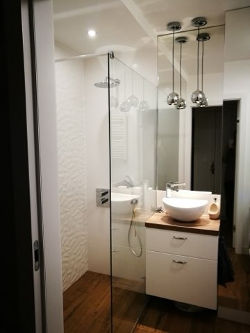 Nowoczesna łazienka z kabiną prysznicową typu walk-in, umywalką nablatową na drewnianym blacie i podłogą z paneli imitujących drewno.