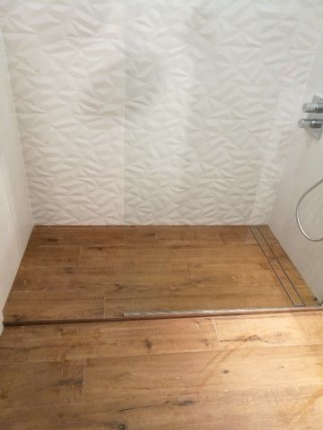 Wykończony brodzik z drewnopodobnych płytek, białe trójwymiarowe płytki ścienne, chromowana bateria prysznicowa.