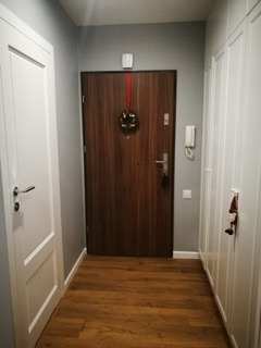 Korytarz po odświeżeniu: drewnopodobna podłoga, szare ściany, białe drzwi i szafa w zabudowie, dekoracyjny wieniec na drzwiach wejściowych.