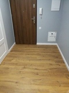 Korytarz po remoncie: jasnobrązowe panele podłogowe imitujące drewno, szare ściany z białymi listwami przypodłogowymi, brązowe drzwi wejściowe, biała skrzynka elektryczna na ścianie.