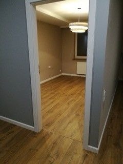 Puste pomieszczenie po remoncie: świeżo ułożona podłoga z paneli w odcieniach drewna, szare ściany, widoczny fragment okna z grzejnikiem i nowoczesna lampa sufitowa. Widok z perspektywy wejścia...