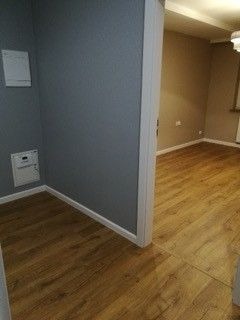 Szare ściany w rogu pokoju z widocznymi skrzynkami elektrycznymi i nową podłogą z paneli laminowanych w odcieniach drewna, widok na sąsiednie pomieszczenie.