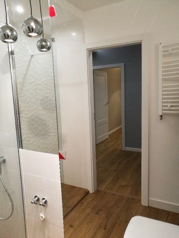 Nowoczesna łazienka z kabiną prysznicową, chromowanymi lampami wiszącymi i drewnopodobnymi panelami podłogowymi, widok na korytarz z pomalowanymi ścianami na szaro.