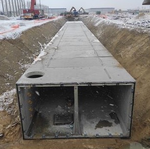 Prefabrykowany betonowy tunel ułożony w wykopie na placu budowy, widoczny żuraw i koparki w tle, częściowo pokryty śniegiem.