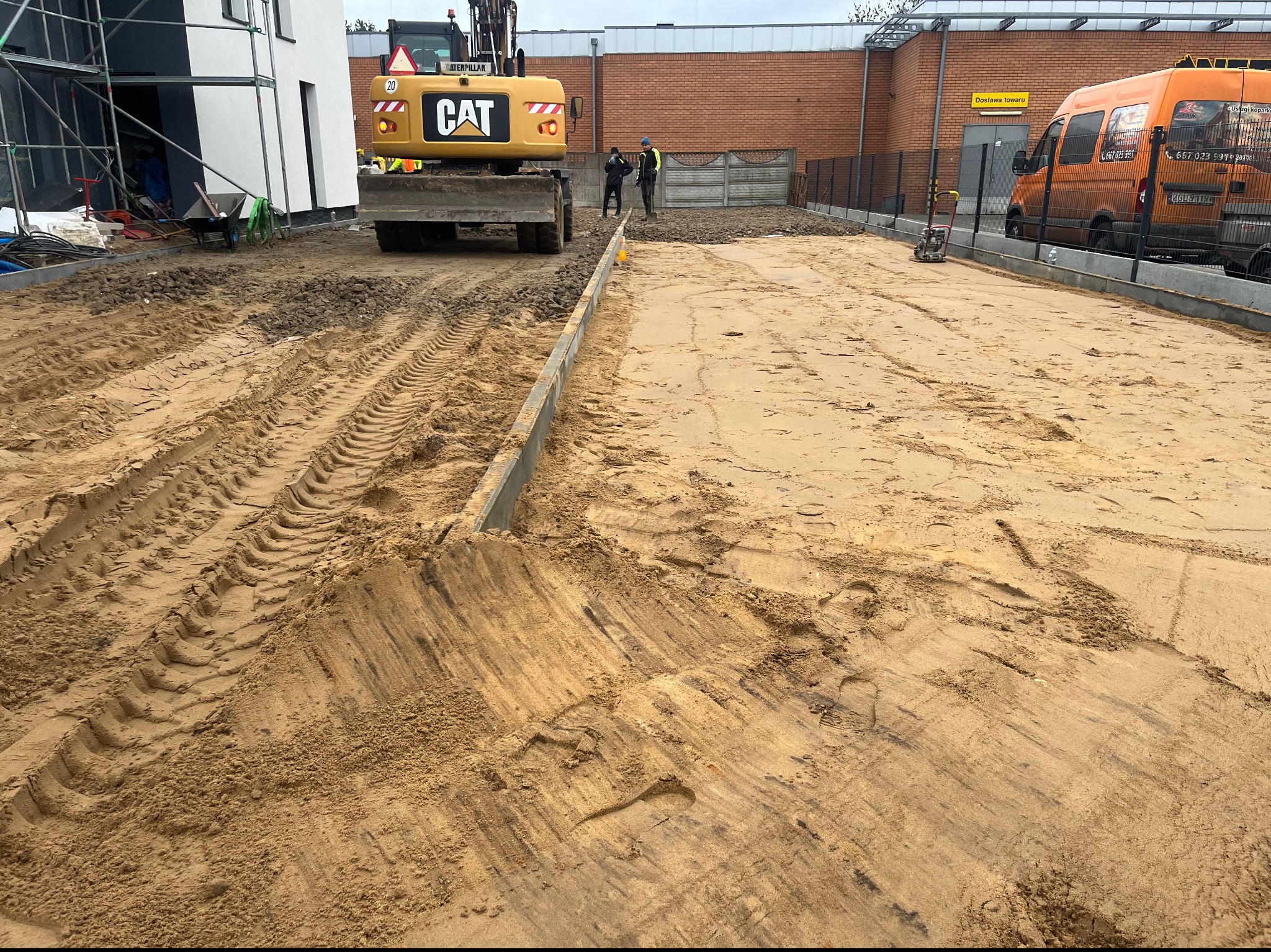 Przygotowanie podłoża pod budowę: wyrównany piasek, krawężniki, koparka CAT i pracownicy w odblaskowych kamizelkach na placu budowy, widoczny budynek w tle.