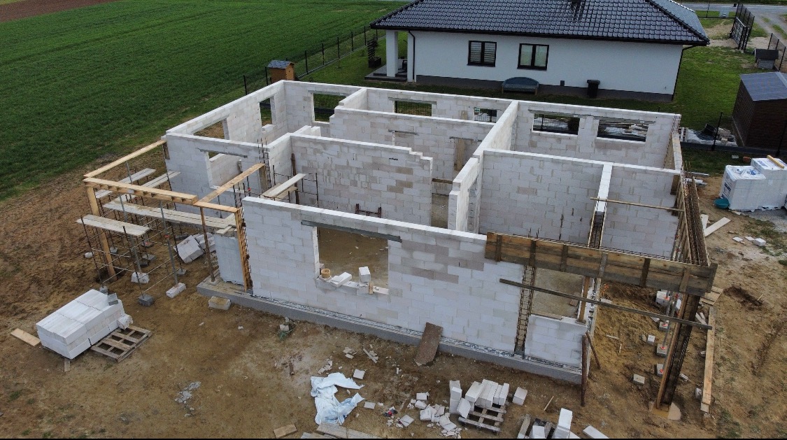 Wznoszenie ścian parterowego domu z białych bloczków, widoczne szalunki i zbrojenia, w tle dom jednorodzinny i zielone pole, ujęcie z drona.
