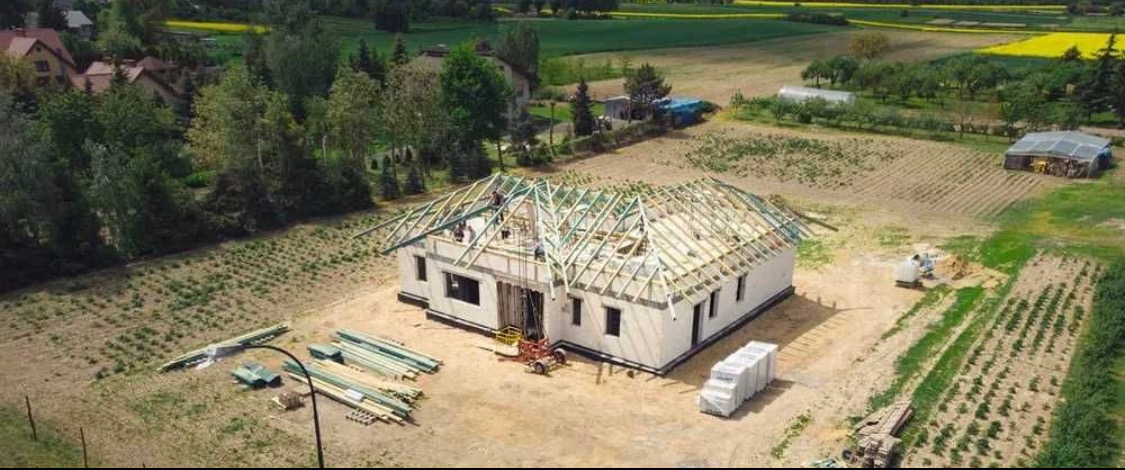 Wznoszenie konstrukcji dachu na parterowym budynku w stanie surowym otwartym, widok z lotu ptaka na plac budowy z przygotowanym drewnem i materiałami izolacyjnymi.