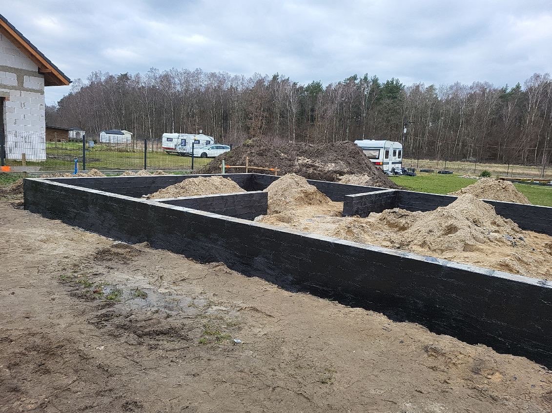 Surowe, czarne fundamenty wylanego domu, z widocznymi pryzmami piasku wewnątrz, na tle zalesionego terenu i przyczep kempingowych.