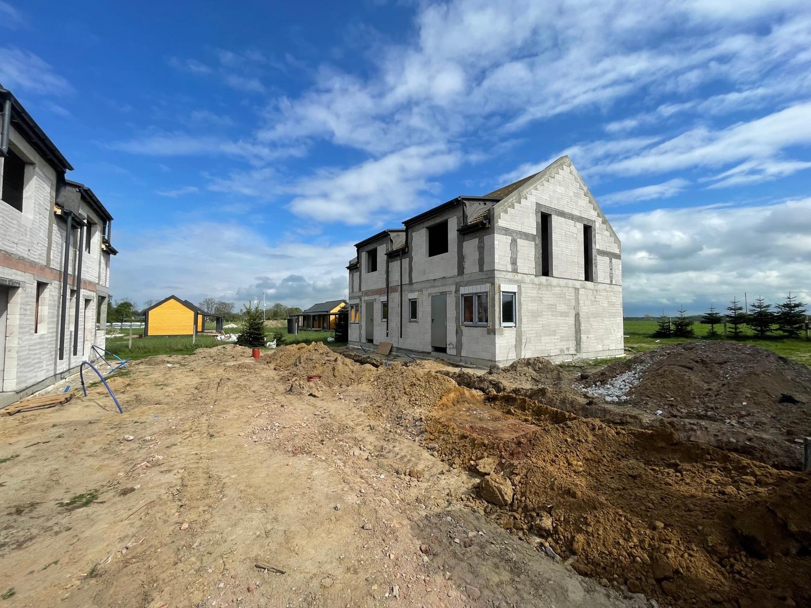 Surowy stan dwóch dwukondygnacyjnych domów z białego bloczka, w tle inne domy i błękitne niebo z chmurami, teren wokół domów rozkopany.