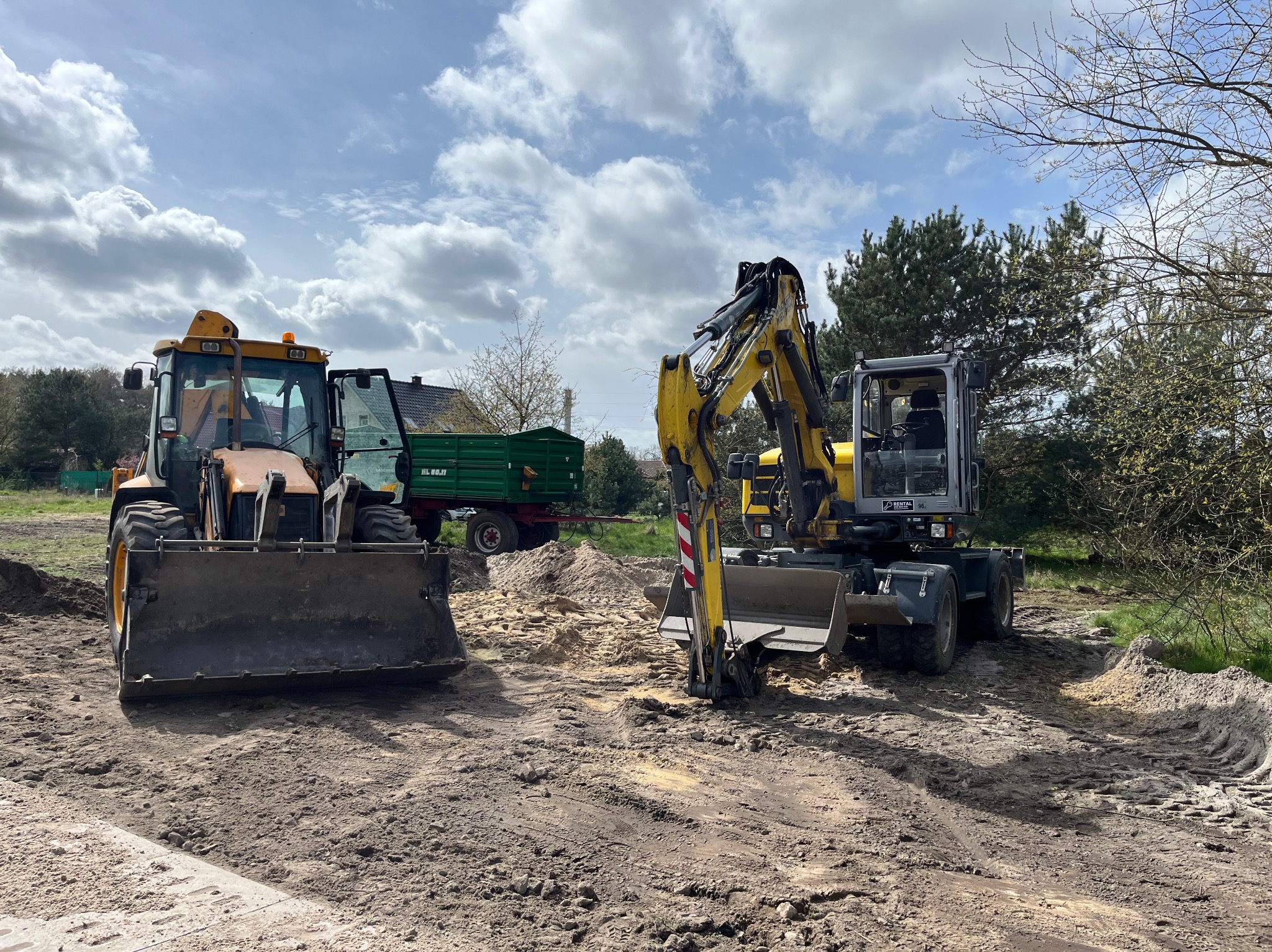 Żółty traktor z otwartą kabiną i szarą łyżką oraz żółta koparka kołowa na placu budowy z hałdami piasku i ziemi, w tle zielona przyczepa i drzewa pod pochmurnym niebem.
