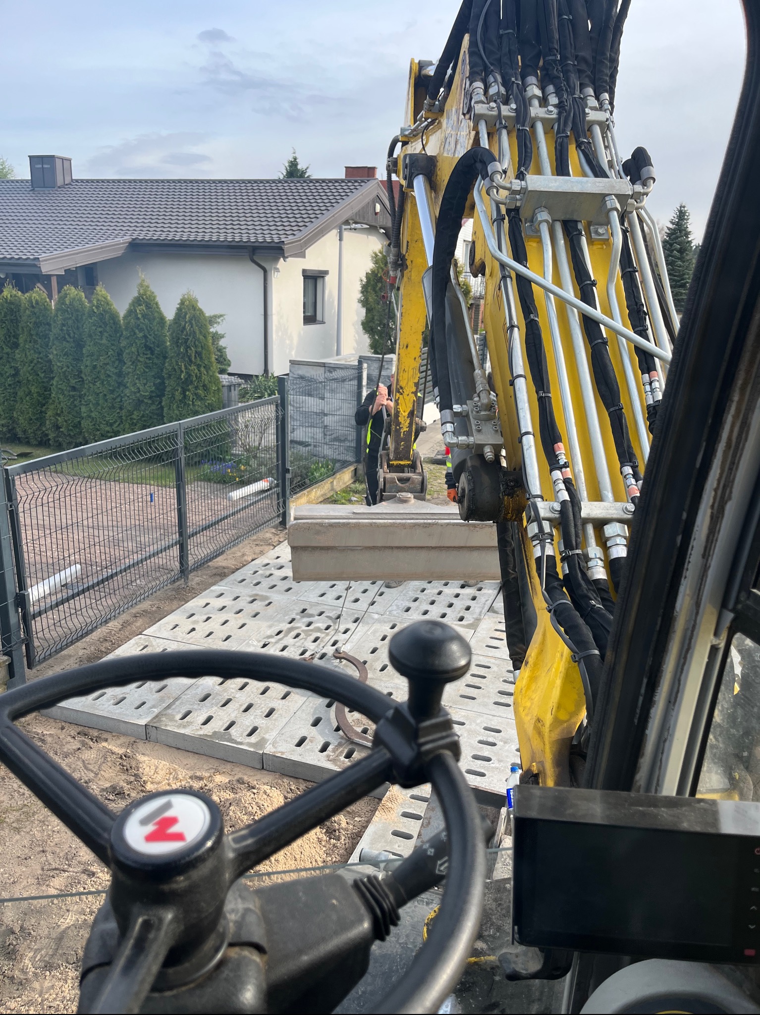 Widok z kabiny żółtej koparki na układanie betonowych płyt drogowych na piasku, w tle ogrodzenie panelowe i dom jednorodzinny.