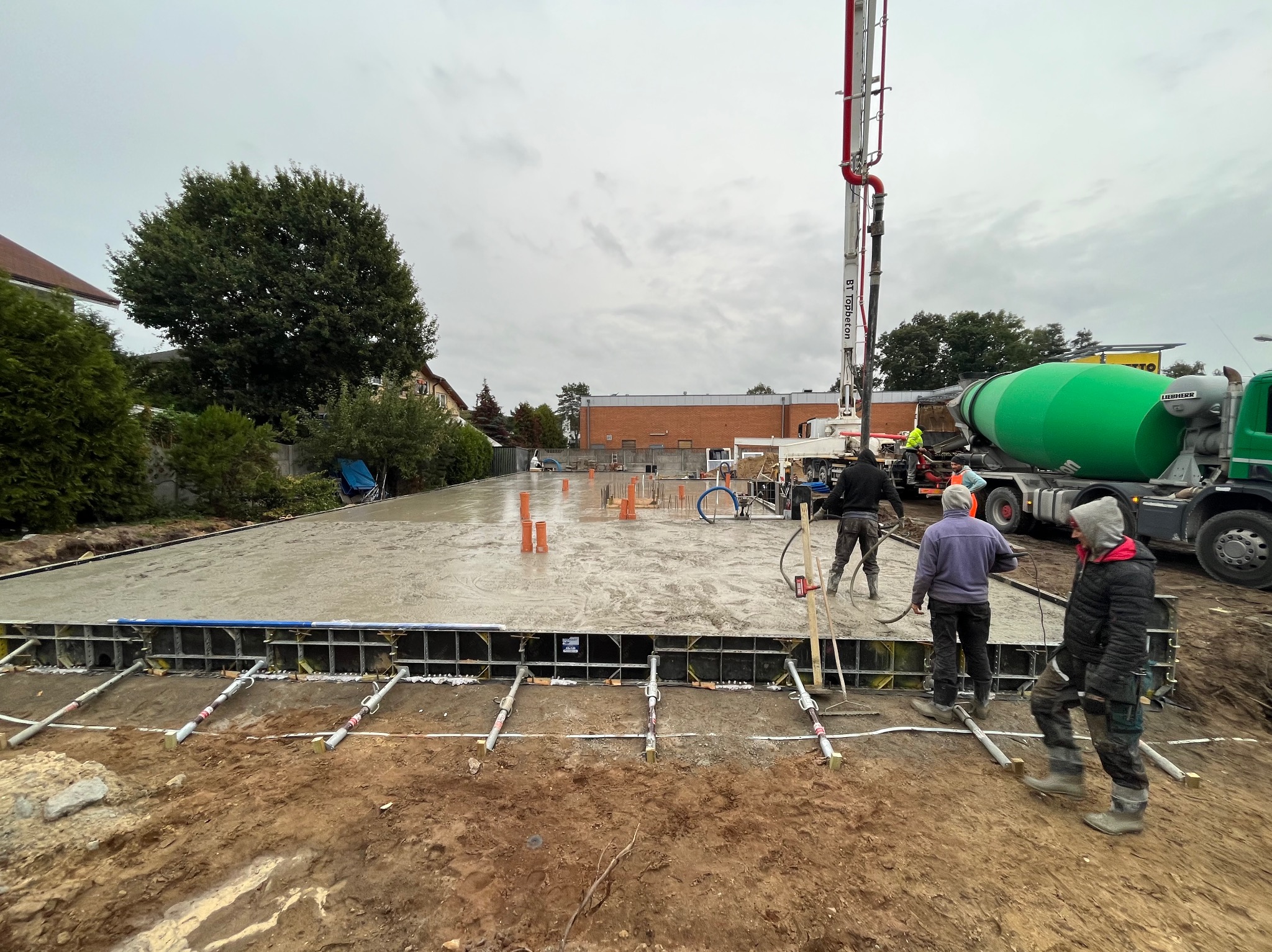 Wylewanie betonu na przygotowany fundament z widocznymi szalunkami, rurami instalacyjnymi i pracownikami obsługującymi pompę do betonu oraz betoniarkę w tle, pochmurny dzień.
