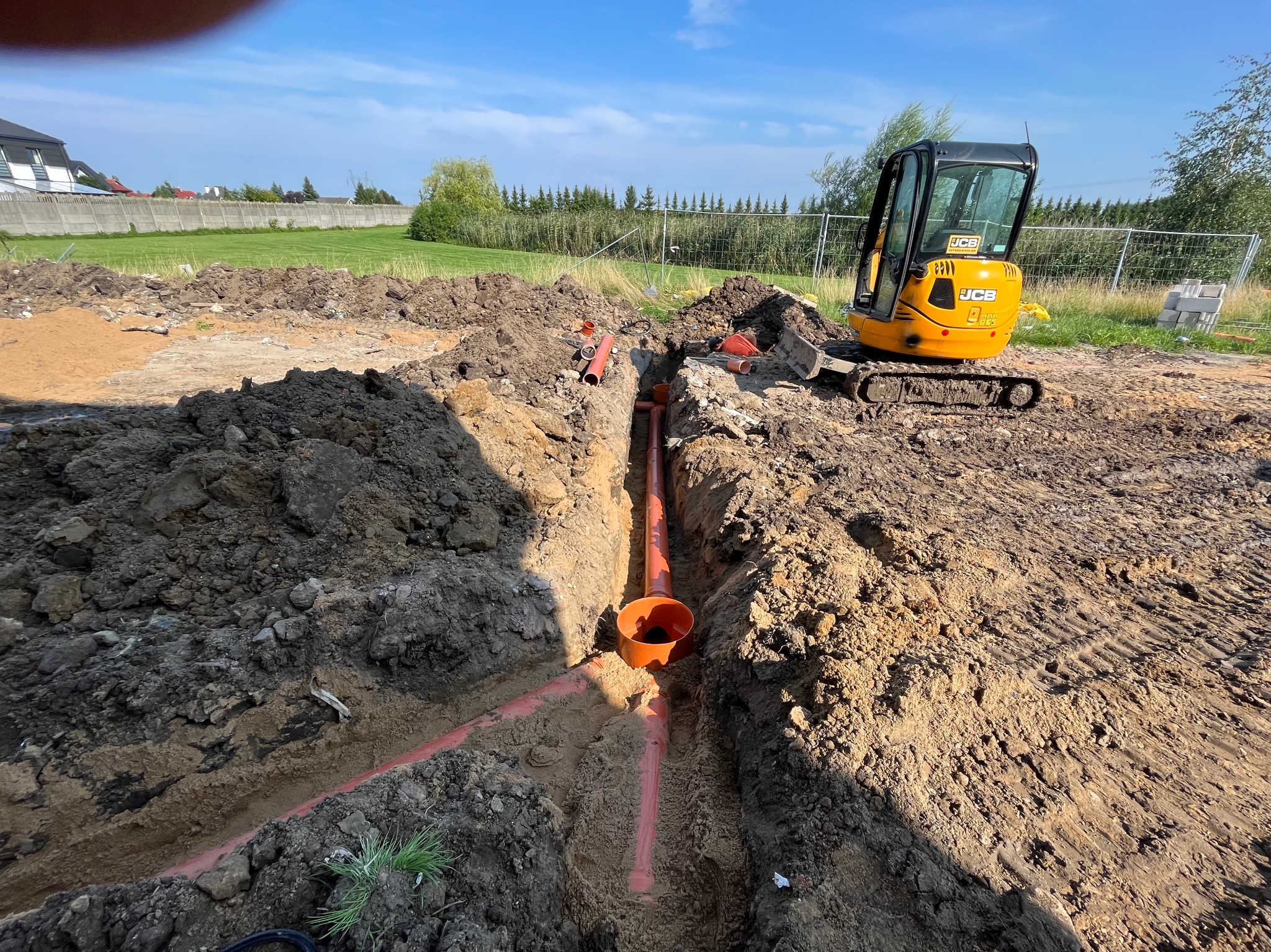 Wykonane wykopy pod rury kanalizacyjne na placu budowy, widoczna mini koparka JCB oraz fragment ogrodzenia w tle.