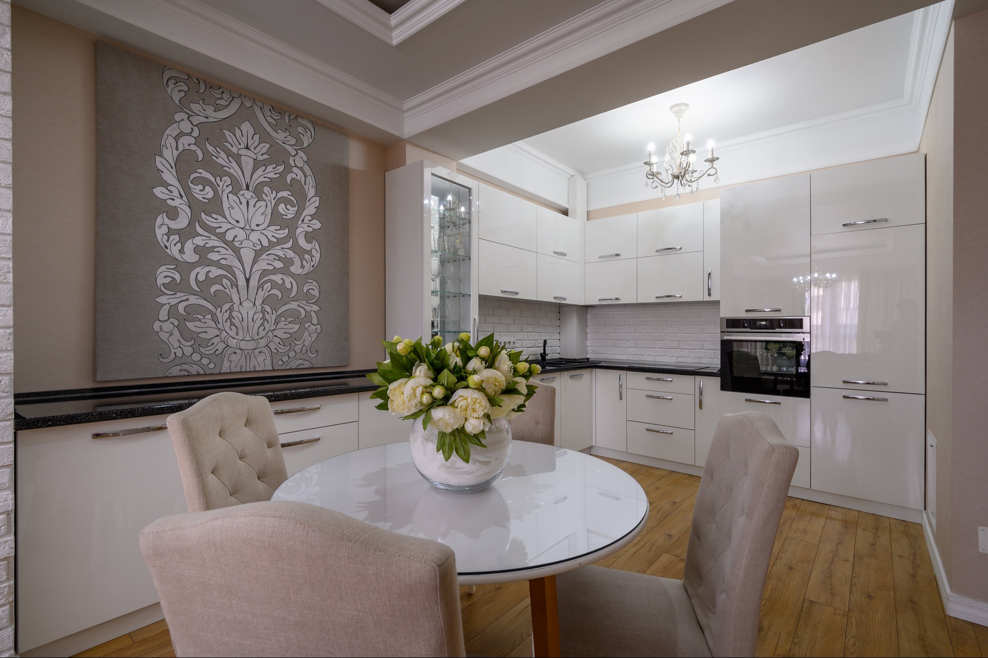 Elegancka, biała kuchnia z połyskiem, okrągły stół z bukietem białych kwiatów, dekoracyjny panel ścienny z ornamentem.