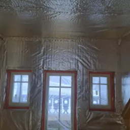 Wnętrze poddasza z trzema oknami w czerwonych ramach, pokryte folią termoizolacyjną, widoczne fragmenty drewnianej konstrukcji dachu.