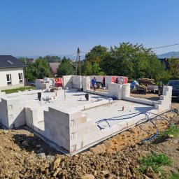 Usługi Ogólnobudowlane SIM-BUD Łukasz Simjak - Budowa domu z bloczków betonowych: fundamenty i pierwsze warstwy ścian w trakcie realizacji, widoczni pracownicy, teren budowy i otaczająca zieleń.