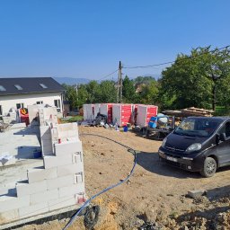 Usługi Ogólnobudowlane SIM-BUD Łukasz Simjak - Budowa domu z bloczków betonu komórkowego: widok na fundamenty, częściowo wzniesione ściany, materiały budowlane, betoniarkę i samochód dostawczy z logo firmy na tle krajobrazu.