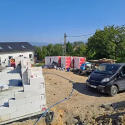 Budowa domu z bloczków betonu komórkowego: widok na fundamenty, częściowo wzniesione ściany, materiały budowlane, betoniarkę i samochód dostawczy z logo firmy na tle krajobrazu.