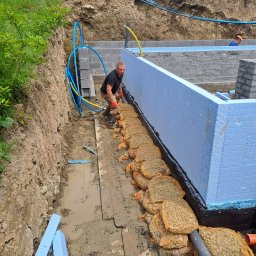 Usługi Ogólnobudowlane SIM-BUD Łukasz Simjak - Budowa fundamentów: Robotnik w brudnych butach układa worki z kruszywem w wykopie obok ściany fundamentowej z izolacją termiczną, w tle widoczna ściana wykopu i drugi pracownik.