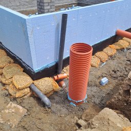 Usługi Ogólnobudowlane SIM-BUD Łukasz Simjak - Izolowane fundamenty z niebieskim styropianem, rury drenażowe i worki z kruszywem, widok na mur z cegieł i pracownika w tle.