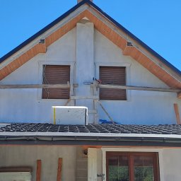 Usługi Ogólnobudowlane SIM-BUD Łukasz Simjak - Fragment elewacji domu w trakcie budowy z widocznym dachem z czarnej dachówki, oknami z roletami, kominem i drewnianymi elementami wykończeniowymi pod dachem; na dachu leżą materiały budowlane...