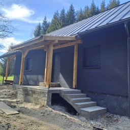 Usługi Ogólnobudowlane SIM-BUD Łukasz Simjak - Nowoczesny dom z ciemną elewacją i metalowym dachem, w trakcie budowy werandy z surowego drewna i betonowych schodów wejściowych, widoczne materiały budowlane na gruncie.