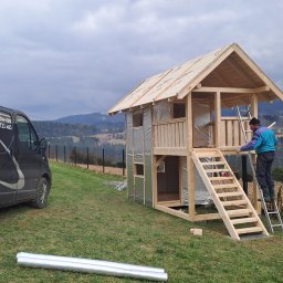 Usługi Ogólnobudowlane SIM-BUD Łukasz Simjak - Budowa dwupoziomowego domku z drewna z izolacją, pracownik na drabinie, w tle górzysty krajobraz i samochód firmy budowlanej.