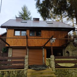 Usługi Ogólnobudowlane SIM-BUD Łukasz Simjak - Dwukondygnacyjny dom z drewnianą elewacją i brązowym, metalowym dachem. Kamienna podmurówka i elementy dekoracyjne z kamienia. Częściowo zasłonięte rolety zewnętrzne.