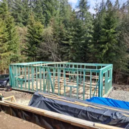 Wznoszenie szkieletu domu na fundamencie, widoczne drewniane elementy konstrukcyjne pomalowane na zielono, w tle las i zaparkowany samochód.