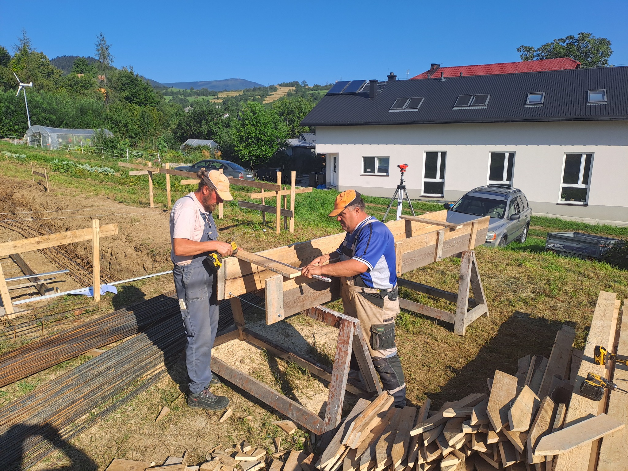 Dwóch budowlańców mierzy deskę na drewnianym szalunku fundamentów domu na tle zielonego wzgórza i domu z panelami słonecznymi na dachu.