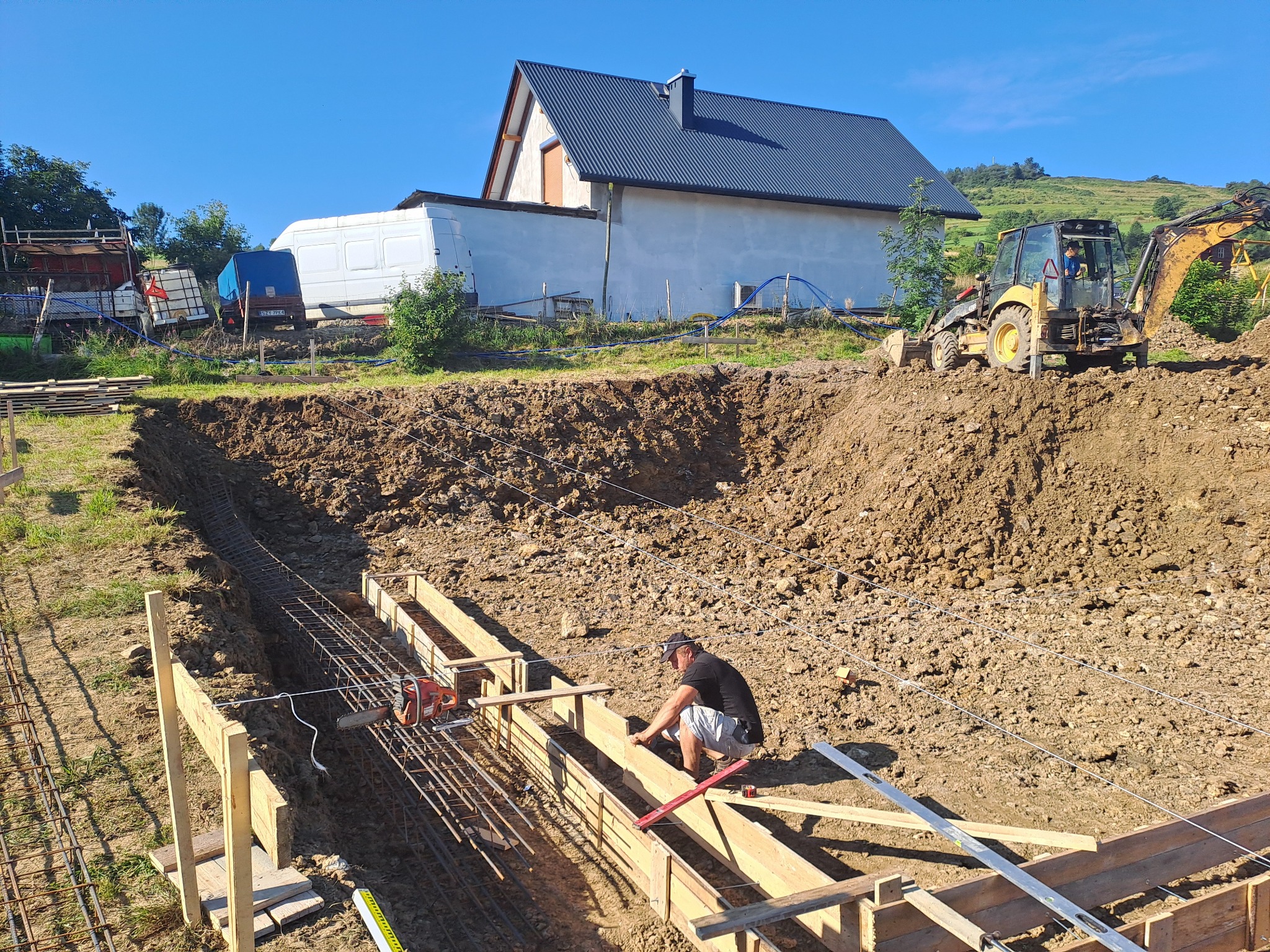Przygotowanie szalunku i zbrojenia pod fundamenty w Soblówce, z widocznym pracownikiem, koparką i budynkiem w tle.