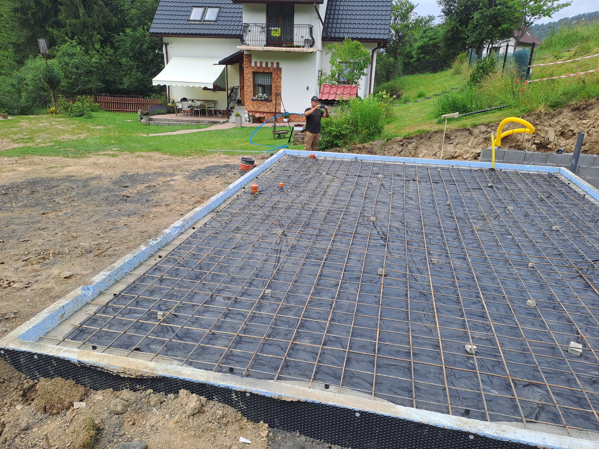Przygotowany zbrojony fundament pod budynek jednorodzinny, z widoczną siatką zbrojeniową na czarnej folii, zabezpieczony niebieskim styropianem, w tle dom z ogrodem i pracownik robiący zdjęcie.