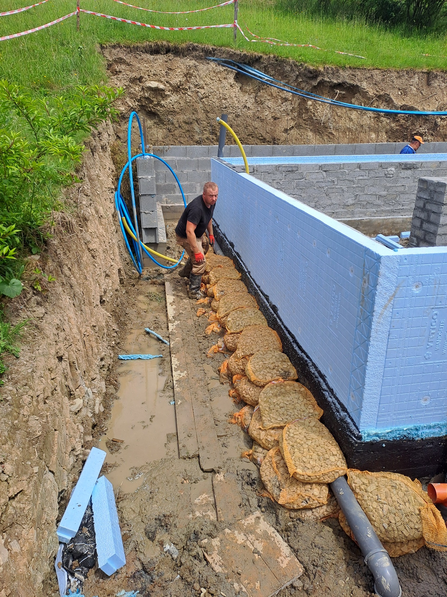 Budowa fundamentów: Robotnik w brudnych butach układa worki z kruszywem w wykopie obok ściany fundamentowej z izolacją termiczną, w tle widoczna ściana wykopu i drugi pracownik.