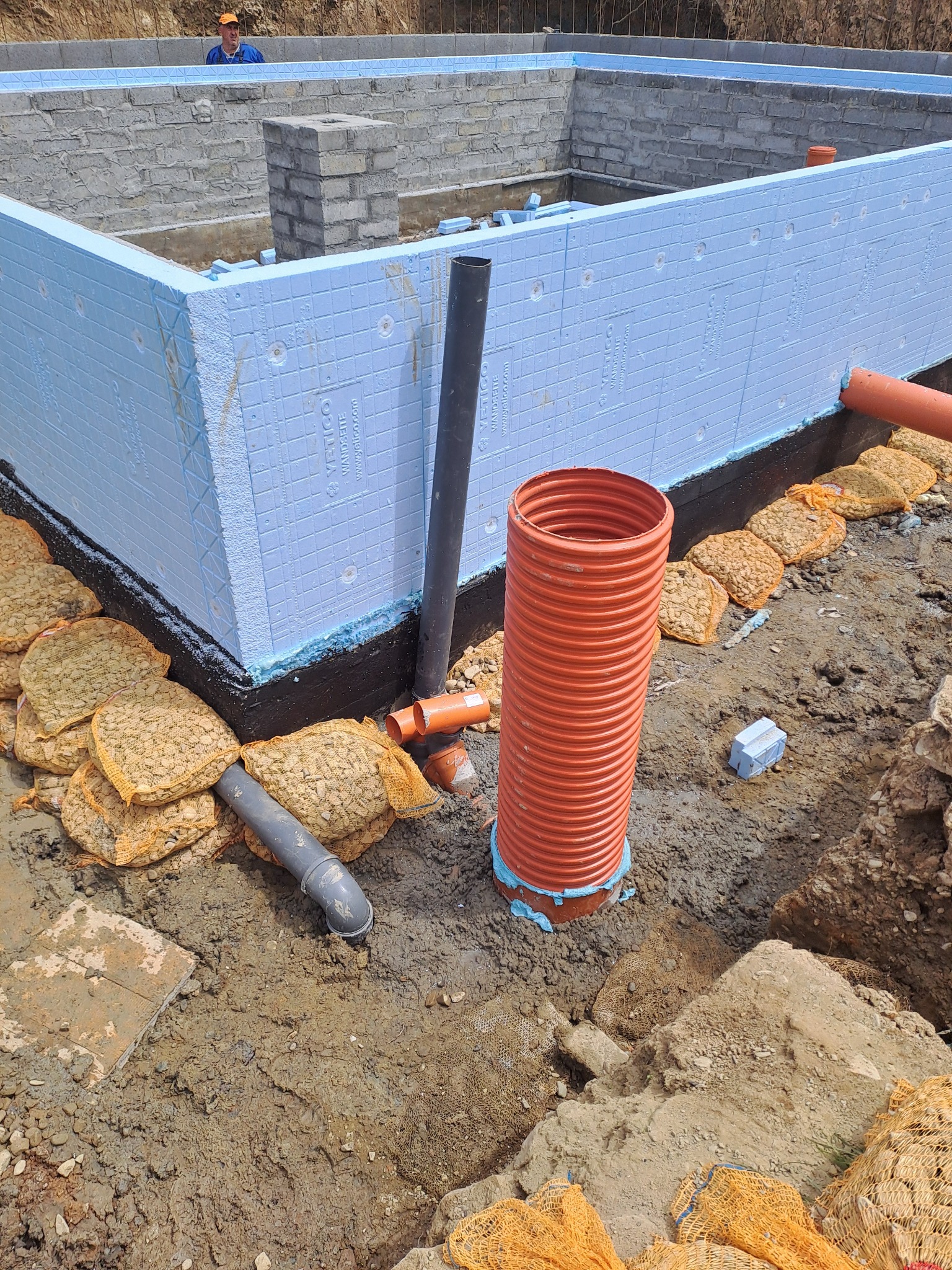 Izolowane fundamenty z niebieskim styropianem, rury drenażowe i worki z kruszywem, widok na mur z cegieł i pracownika w tle.