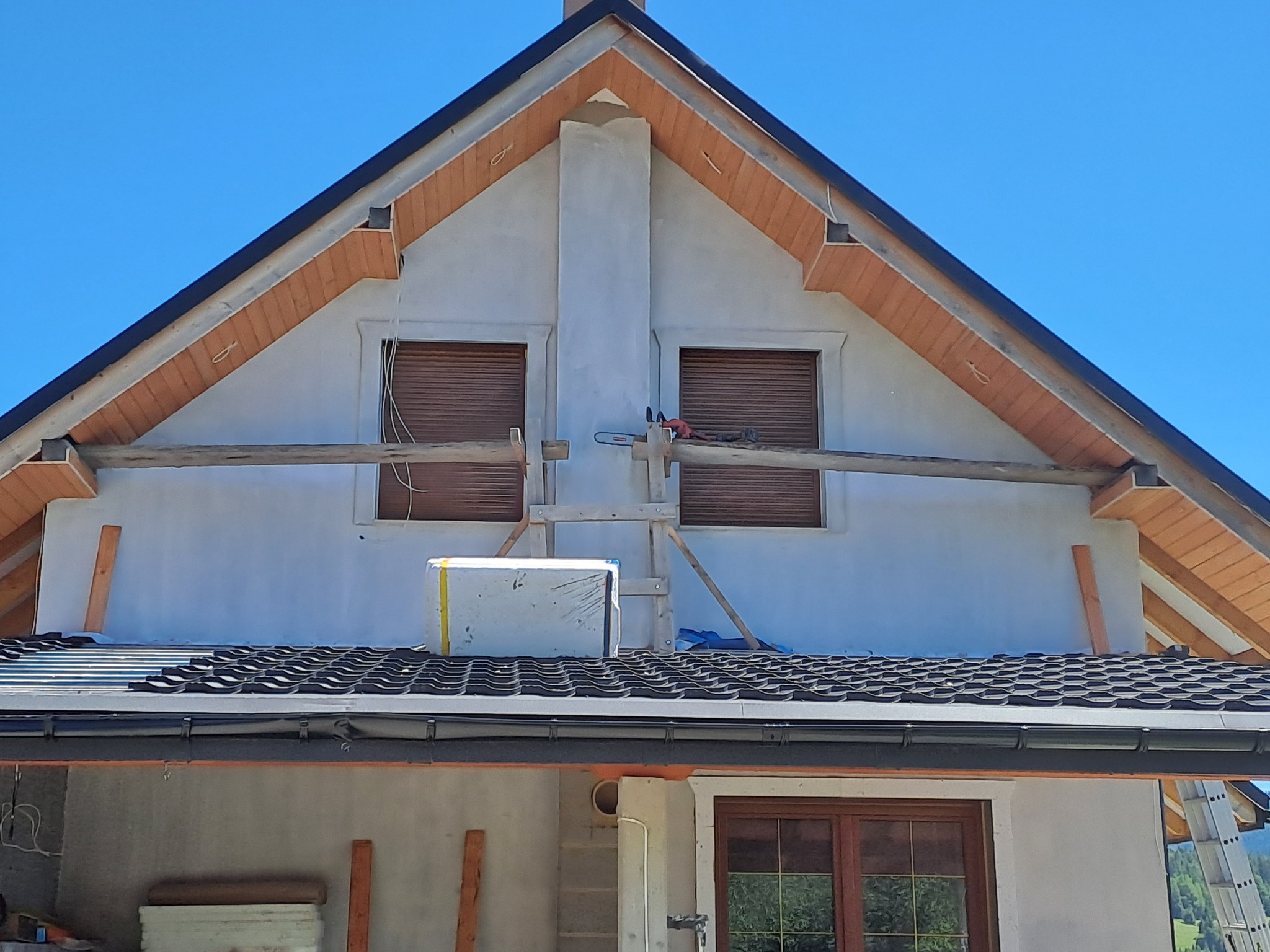 Fragment elewacji domu w trakcie budowy z widocznym dachem z czarnej dachówki, oknami z roletami, kominem i drewnianymi elementami wykończeniowymi pod dachem; na dachu leżą materiały budowlane...