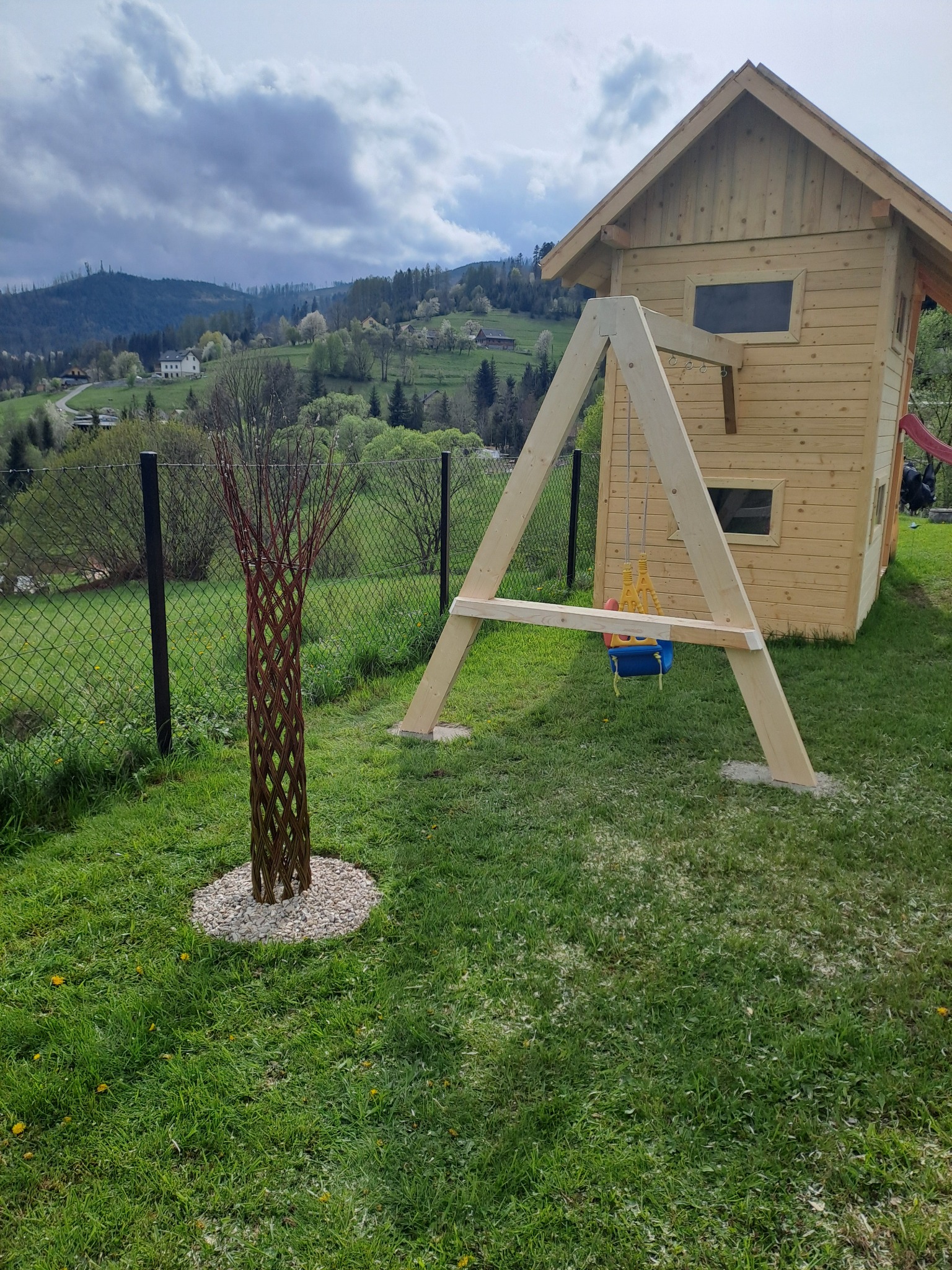 Drewniany domek zabaw dla dzieci z huśtawką, umieszczony na trawniku z widokiem na zalesione wzgórza w tle, obok ozdobna plecionka z gałęzi na kamiennym podłożu.