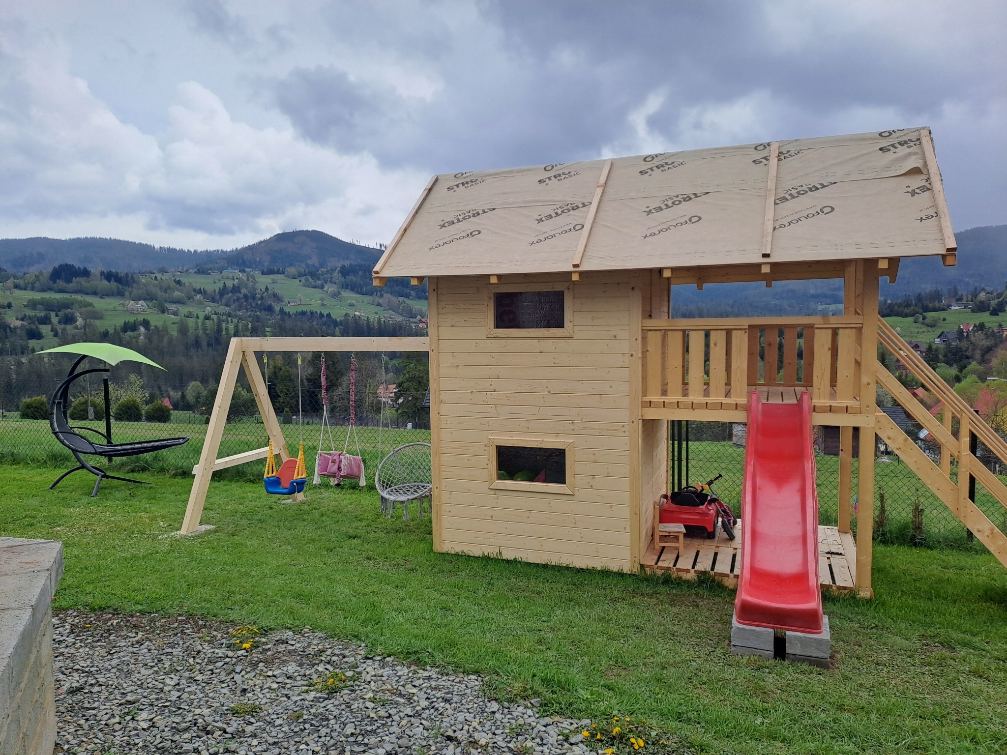Drewniany plac zabaw z domkiem, zjeżdżalnią i huśtawkami, częściowo zadaszony membraną dachową, na tle zielonego trawnika i górskiego krajobrazu.