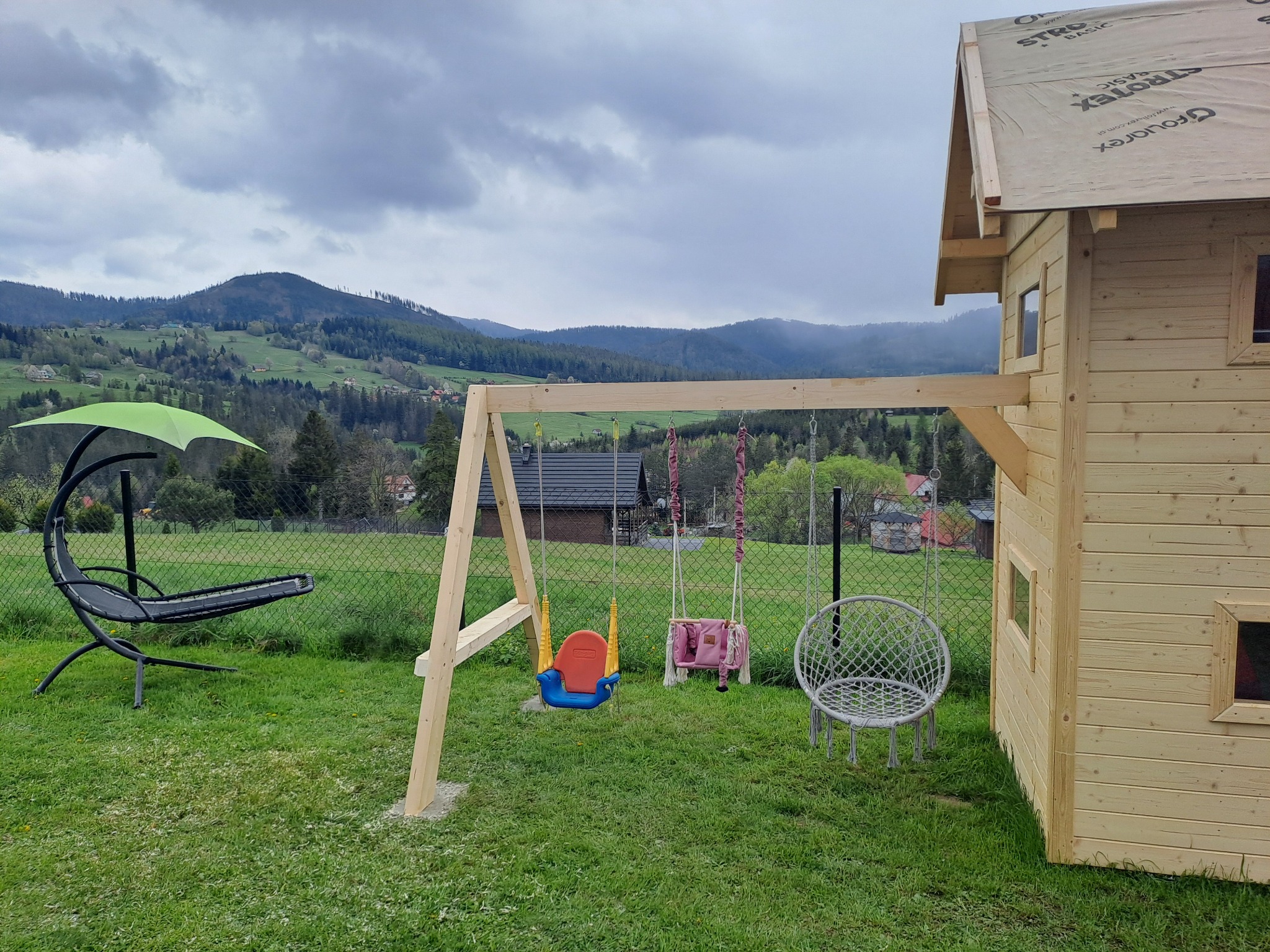 Niedokończony drewniany domek dla dzieci z huśtawkami na tle górskiego krajobrazu w pochmurny dzień, widoczne materiały budowlane na dachu.