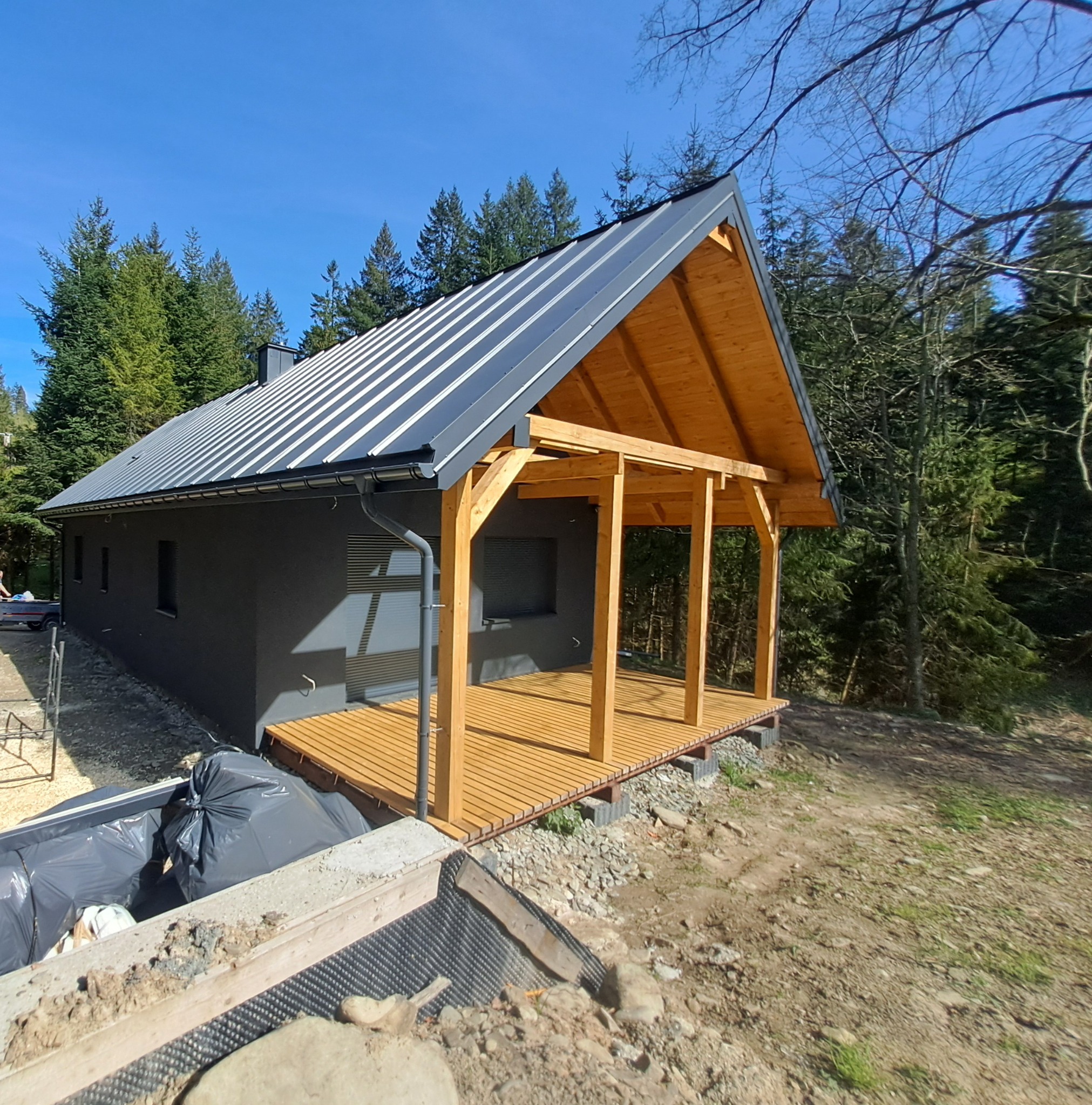 Nowoczesny dom z ciemną elewacją i metalowym dachem, z drewnianą konstrukcją werandy w surowym stanie, widoczne prace wykończeniowe przy fundamentach.