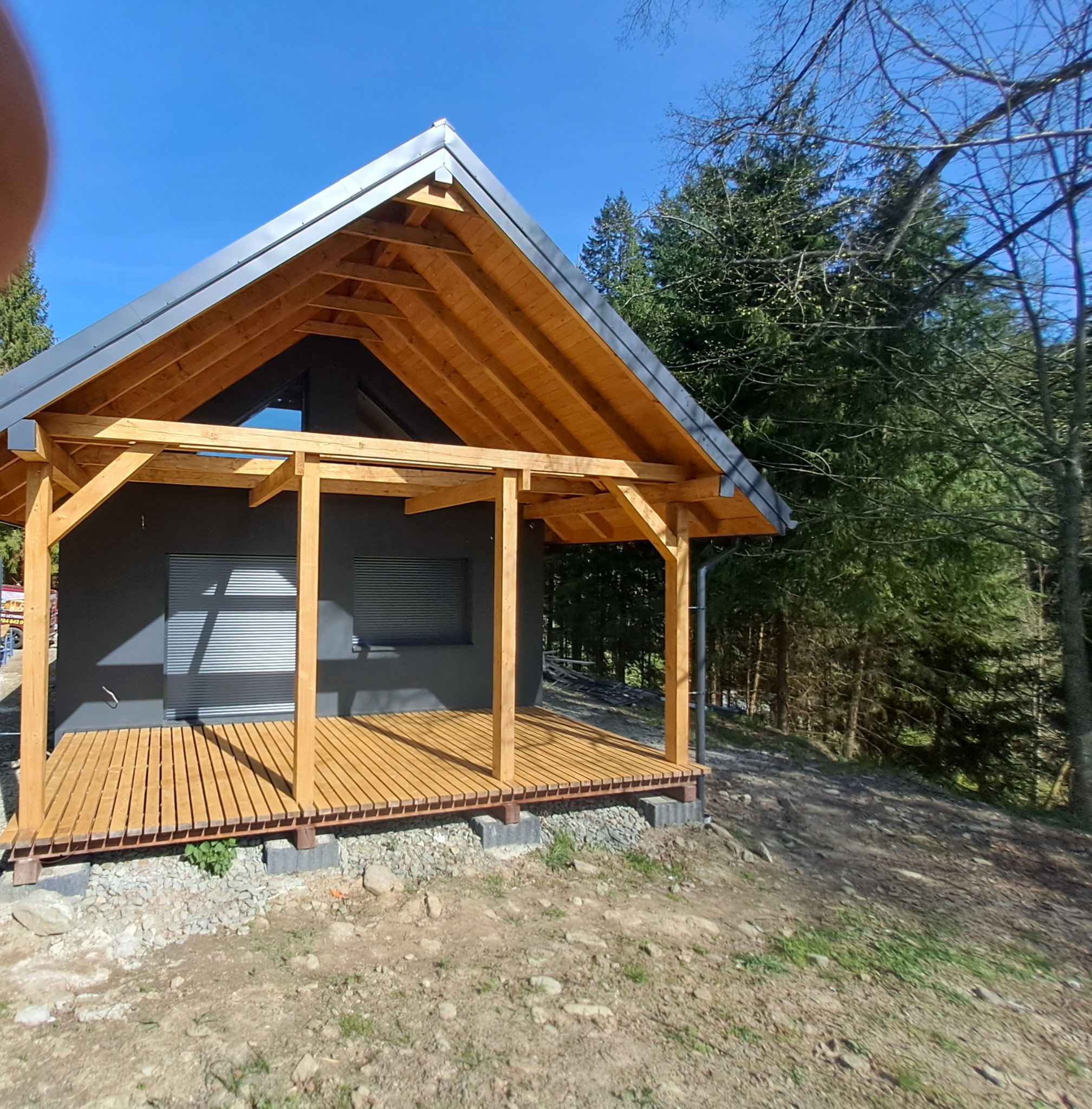 Surowy stan domu z drewnianą konstrukcją nośną, widoczne szare ściany i drewniany taras, otoczony drzewami w słoneczny dzień.