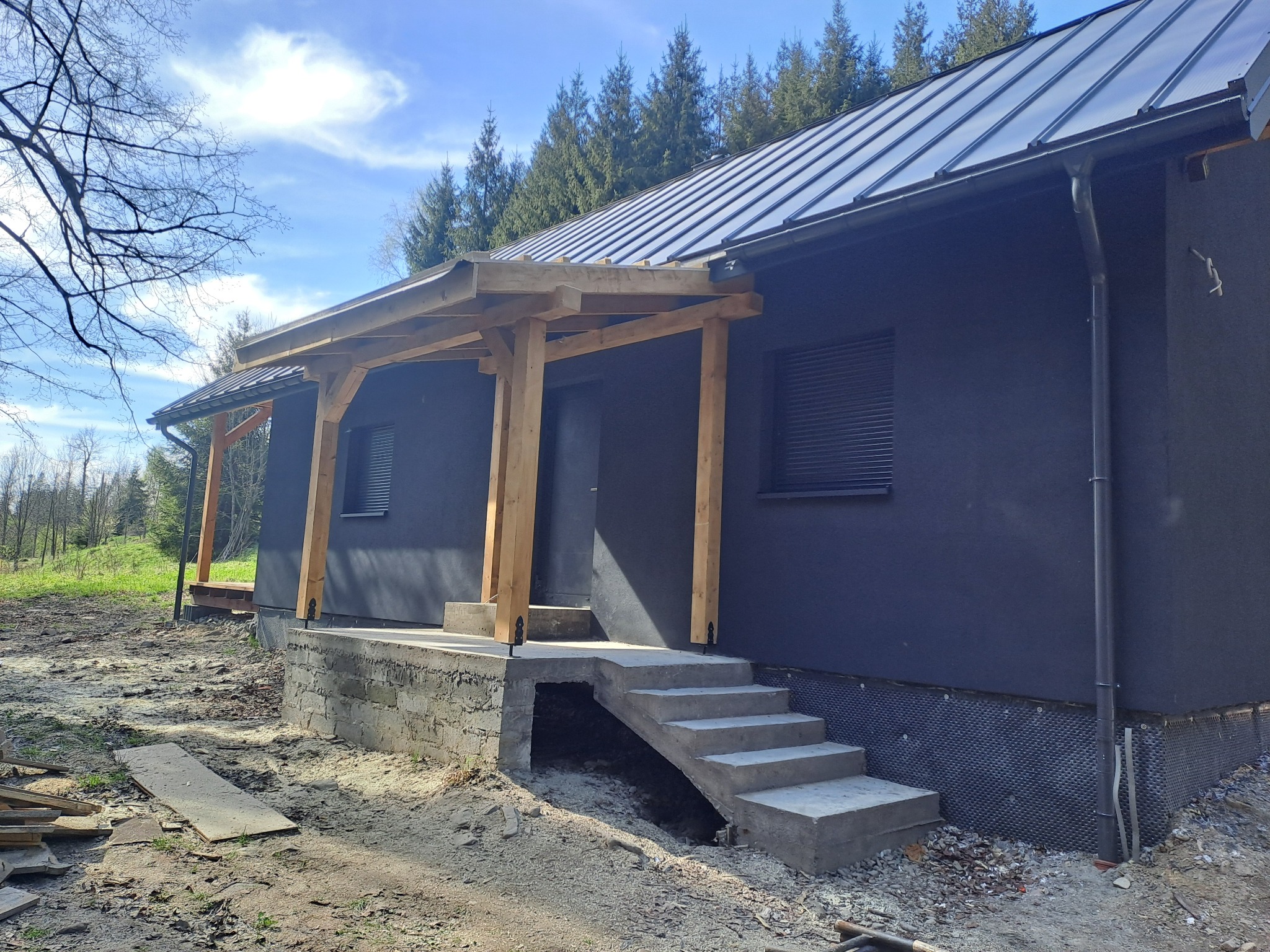 Nowoczesny dom z ciemną elewacją i metalowym dachem, w trakcie budowy werandy z surowego drewna i betonowych schodów wejściowych, widoczne materiały budowlane na gruncie.