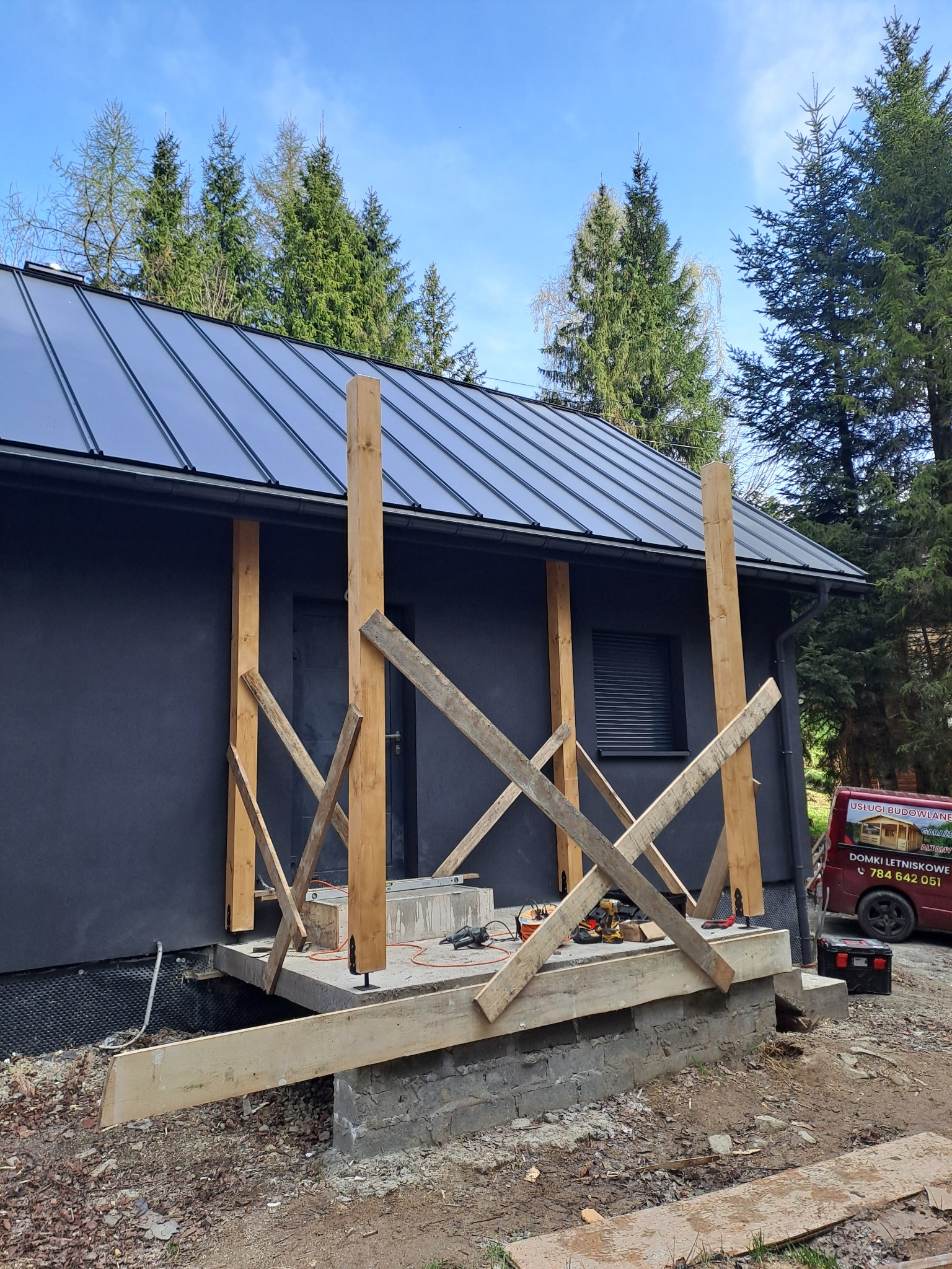 Budynek z ciemną elewacją i metalowym dachem w trakcie budowy werandy z drewnianymi słupami i tymczasowymi belkami, narzędzia budowlane widoczne na podeście.